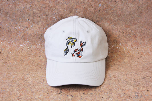 A cream colored dad hat with 2 embroidered koi fish circling on a cork background. One koi fish is gold and grey, and the other is scarlet and grey.