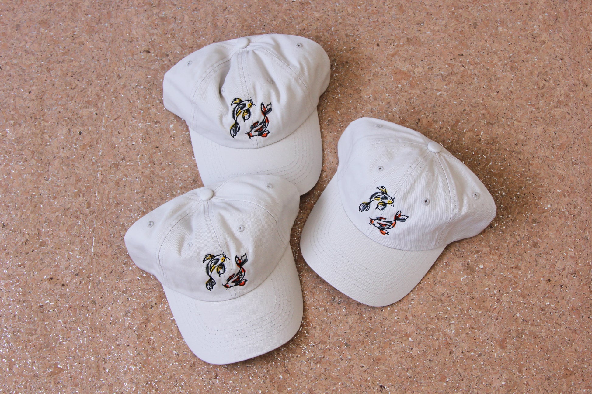 3 cream colored dad hats each with 2 embroidered koi fish circling on a cork background. One koi fish is gold and grey, and the other is scarlet and grey.