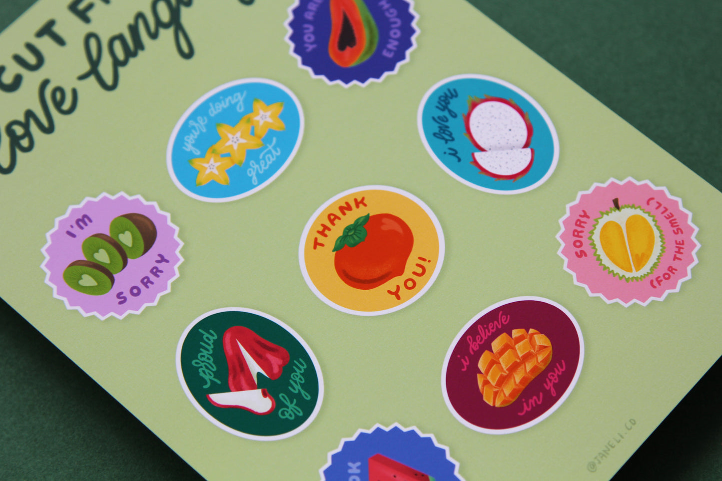A close up of a JaneLi.Co print that says "Cut Fruit Love Language" with 9 different fruit stickers with different messages like "I love you", "I'm sorry", and "I'm proud of you" over a green background.