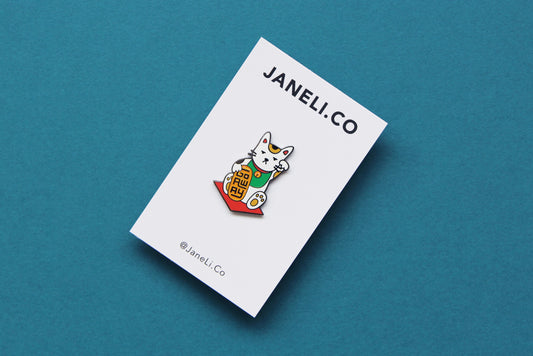 An enamel pin showing maneki neko cat holding a glittery gold bar that says "Go Away" on a white JaneLi.Co backing card over a teal blackground.