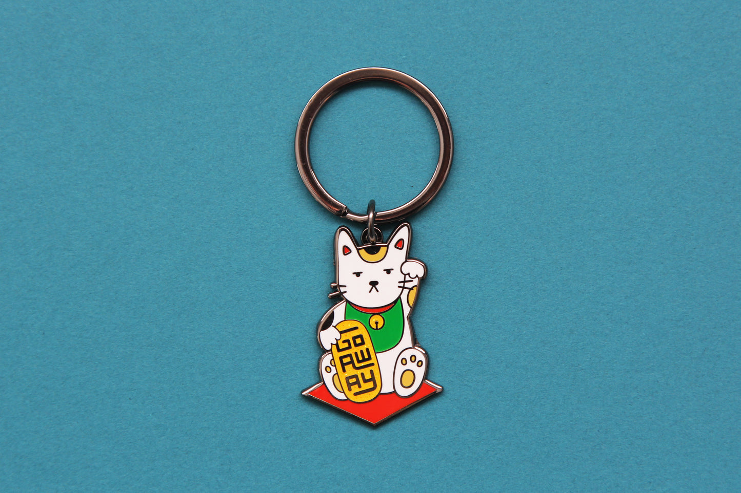 An enamel keychain showing maneki neko cat holding a glittery gold bar that says "Go Away" over a teal background.