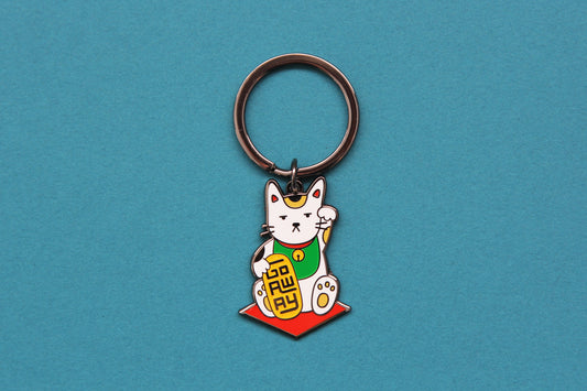 An enamel keychain showing maneki neko cat holding a glittery gold bar that says "Go Away" over a teal background.