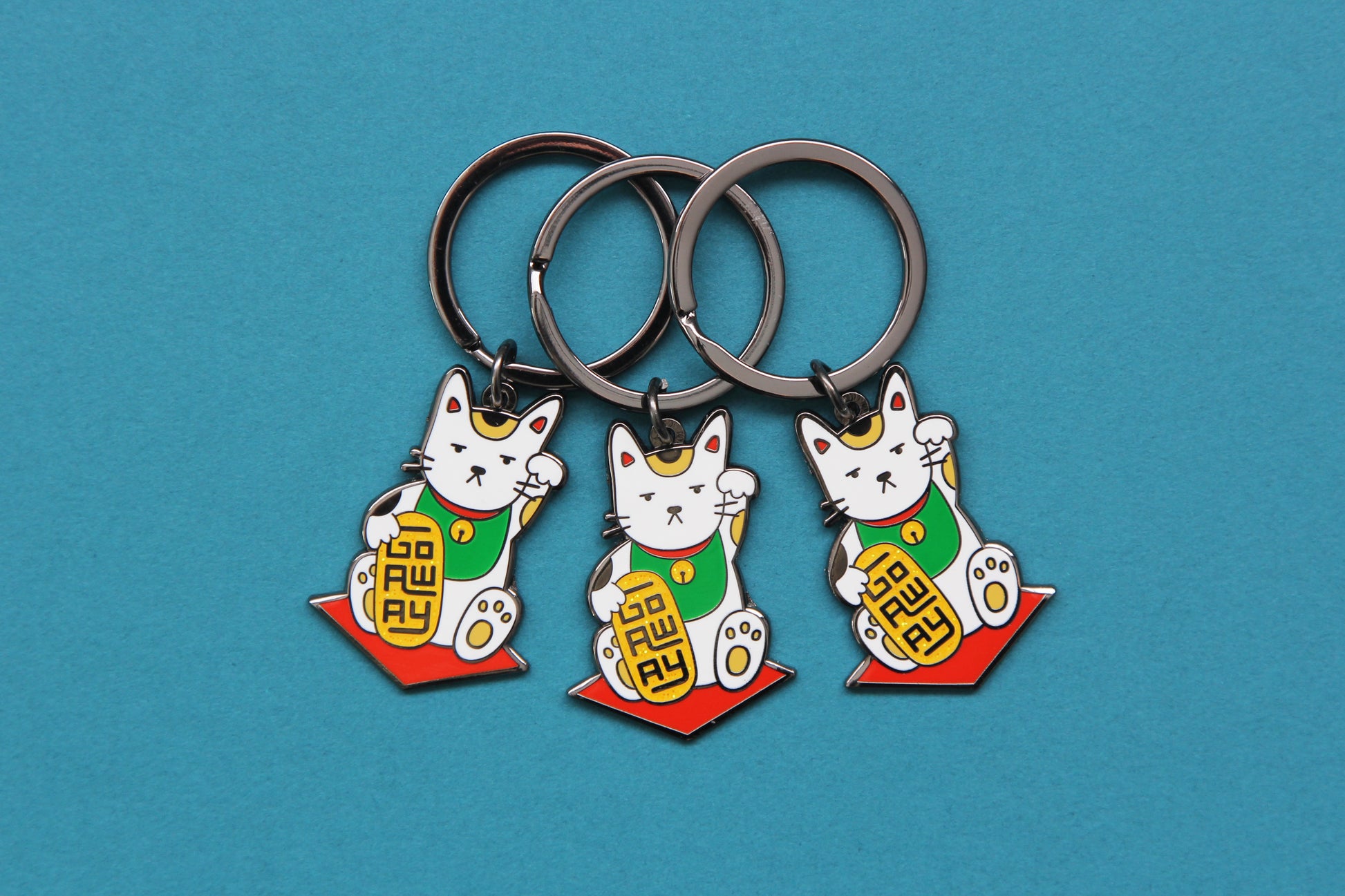 3 enamel keychains showing maneki neko cats holding glittery gold bars that say "Go Away" over a teal background.