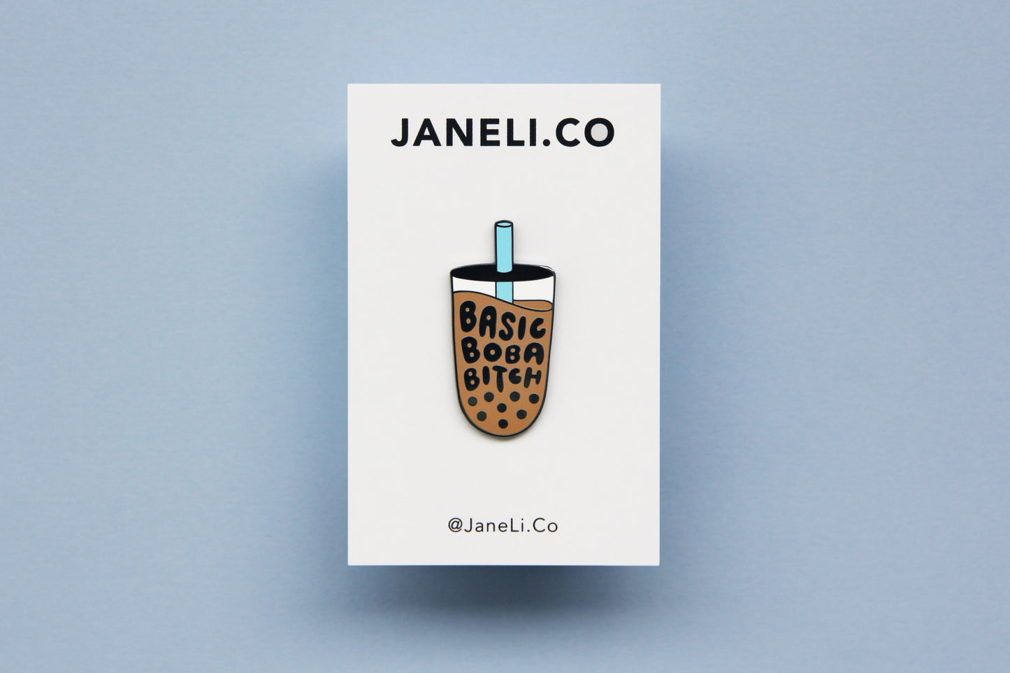An enamel pin showing a cup of boba that says "Basic Boba Bitch" with a blue straw on a white JaneLi.Co backing card over a blue background.