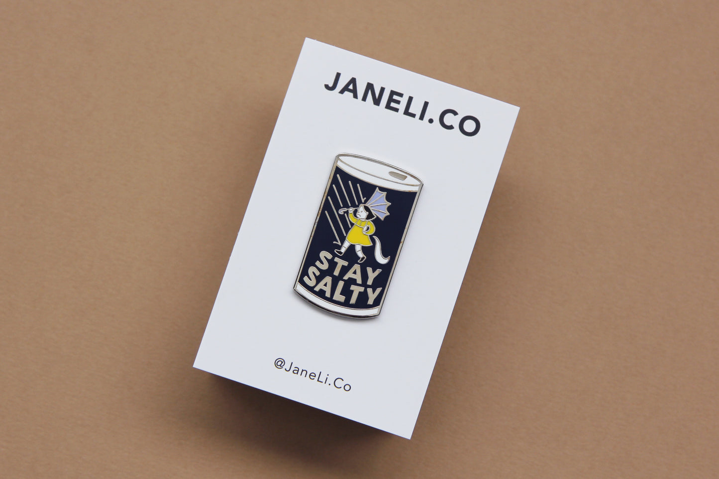 An enamel pin showing a grumpy girl on a salt can that says "Stay Salty" on a white JaneLi.Co backing card over a tan background.