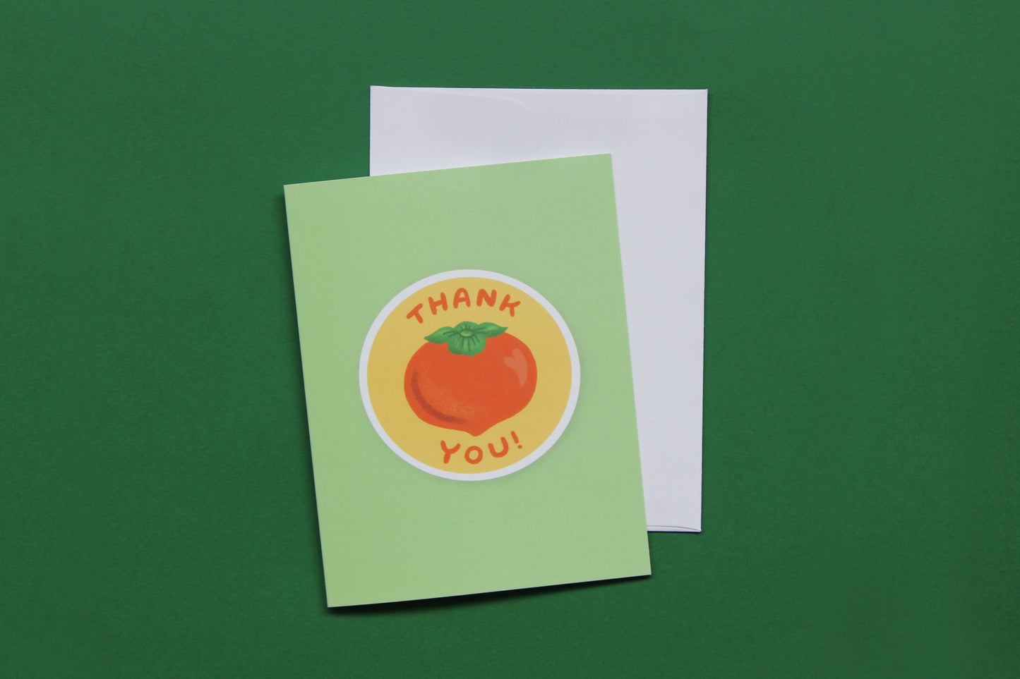 A photo of a green greeting card with a persimmon that says "Thank you" and a white envelope on a green background.