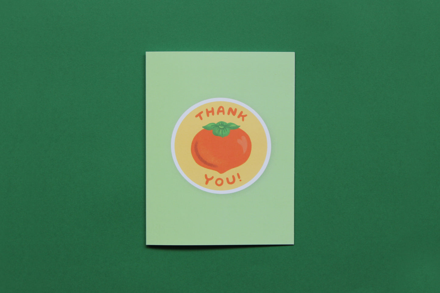 A photo of a green greeting card with a persimmon that says "Thank you" on a green background.