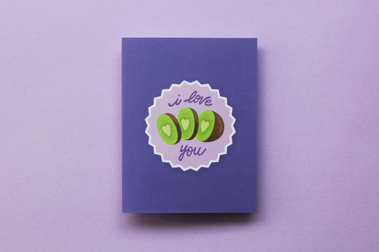 A photo of a lavender greeting card with sliced kiwi that says "I love you" on a purple background.
