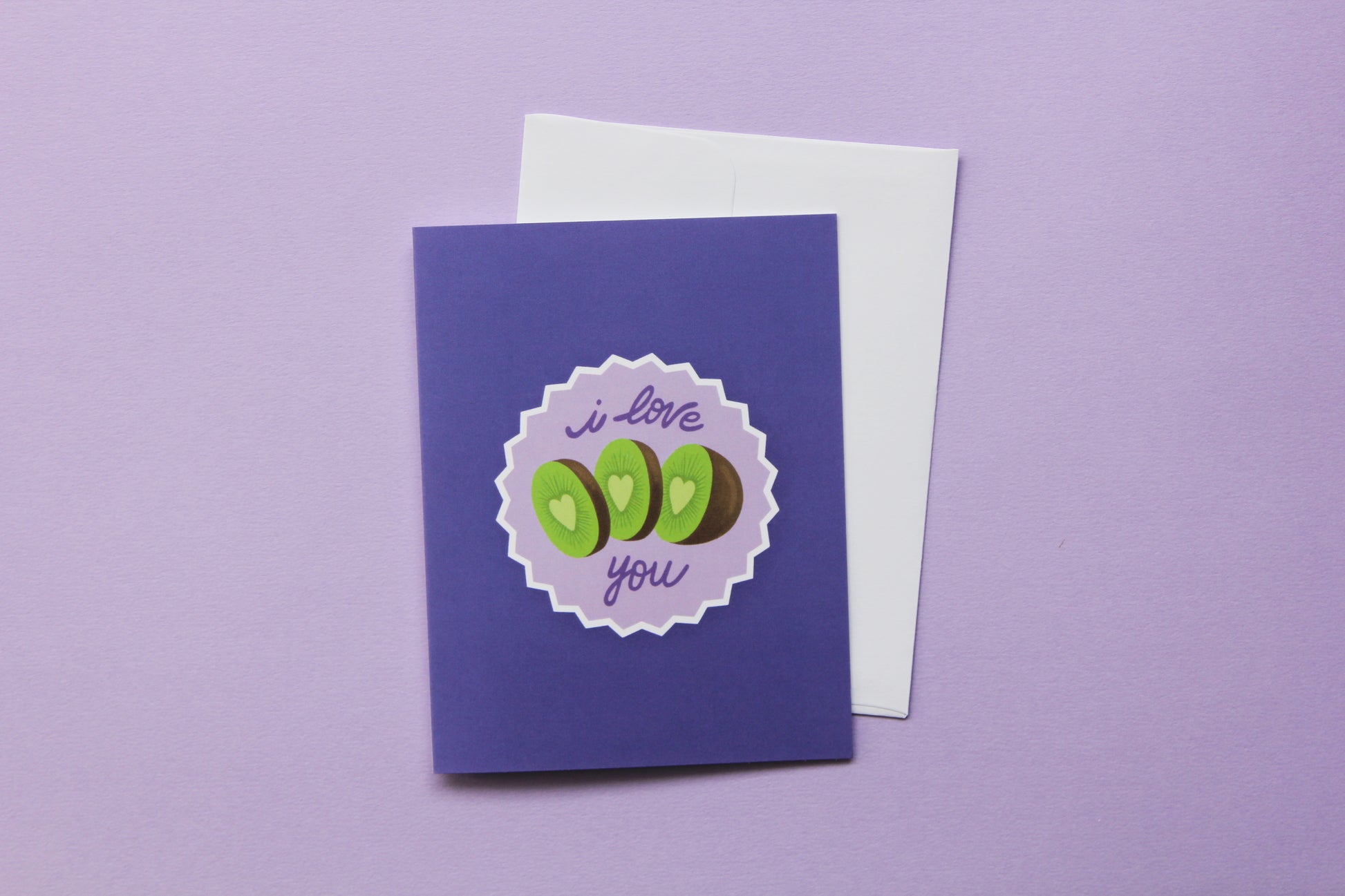 A photo of a lavender greeting card with sliced kiwi that says "I love you" and a white envelope on a purple background.