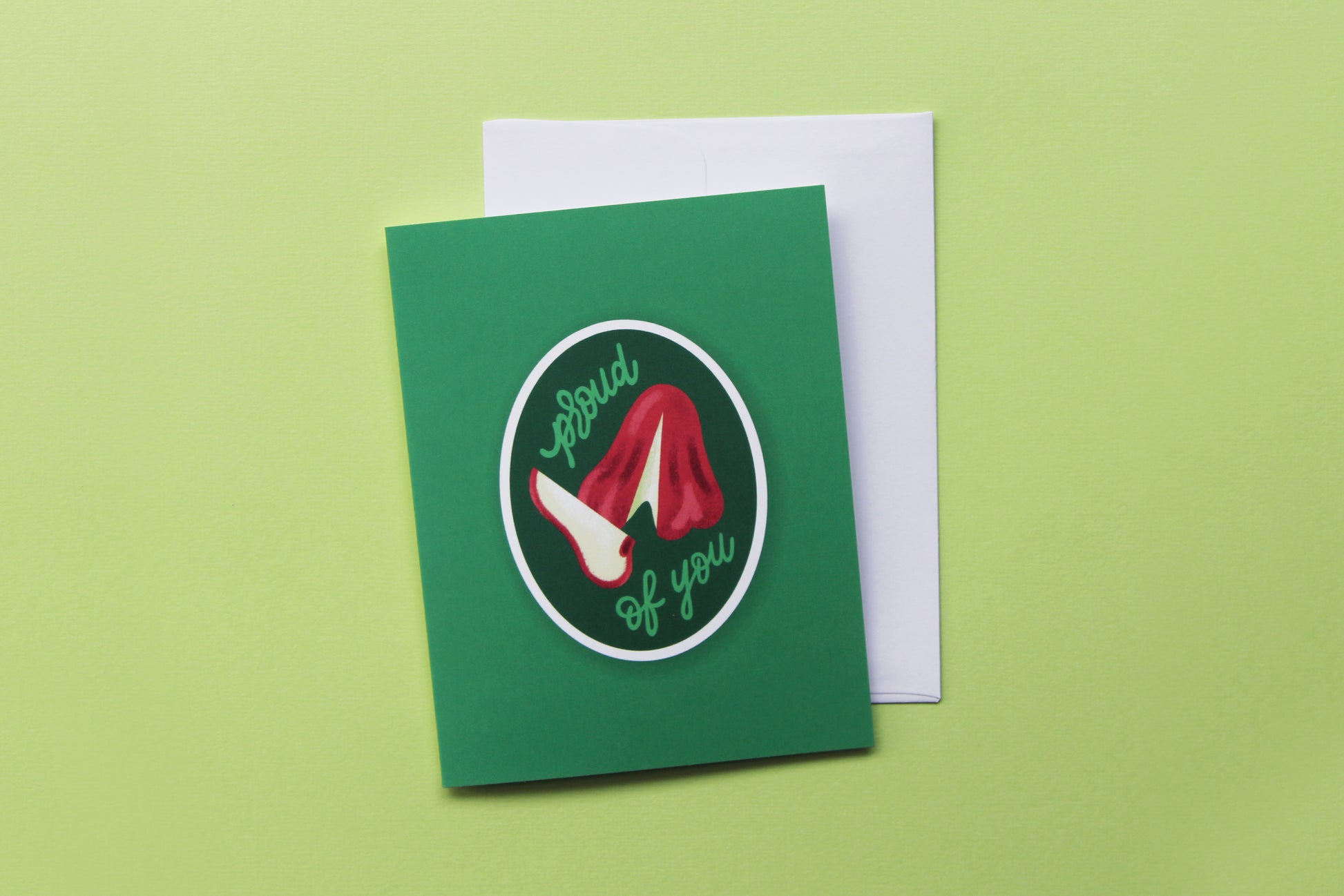 A photo of a green greeting card with a sliced wax apple that says "Proud of you" and a white envelope on a green background.