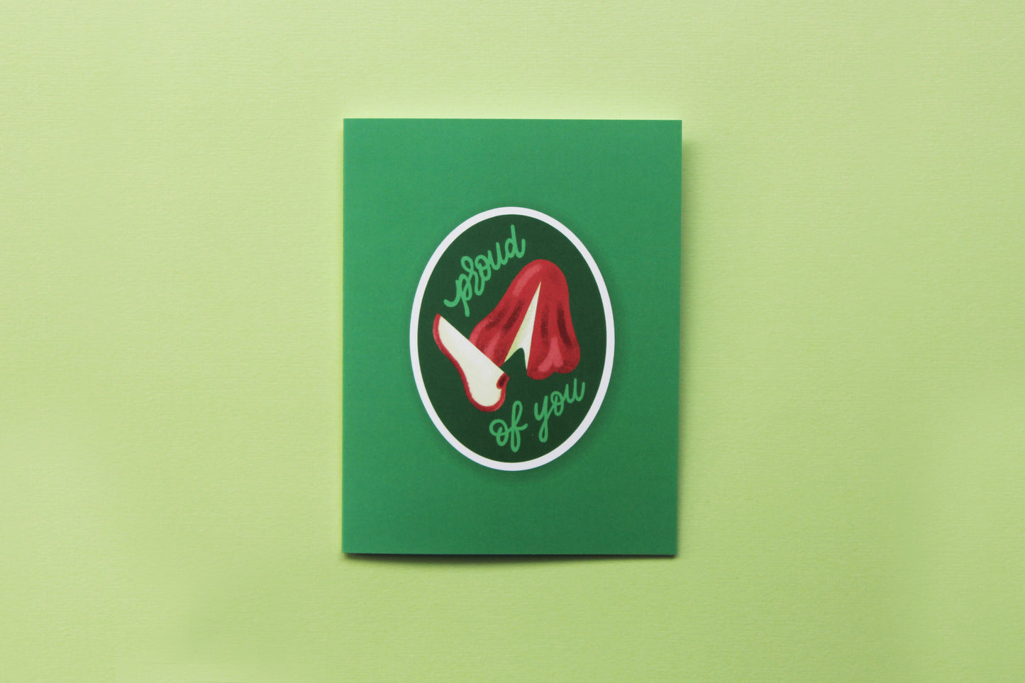 A photo of a green greeting card with a sliced wax apple that says "Proud of you"on a green background.