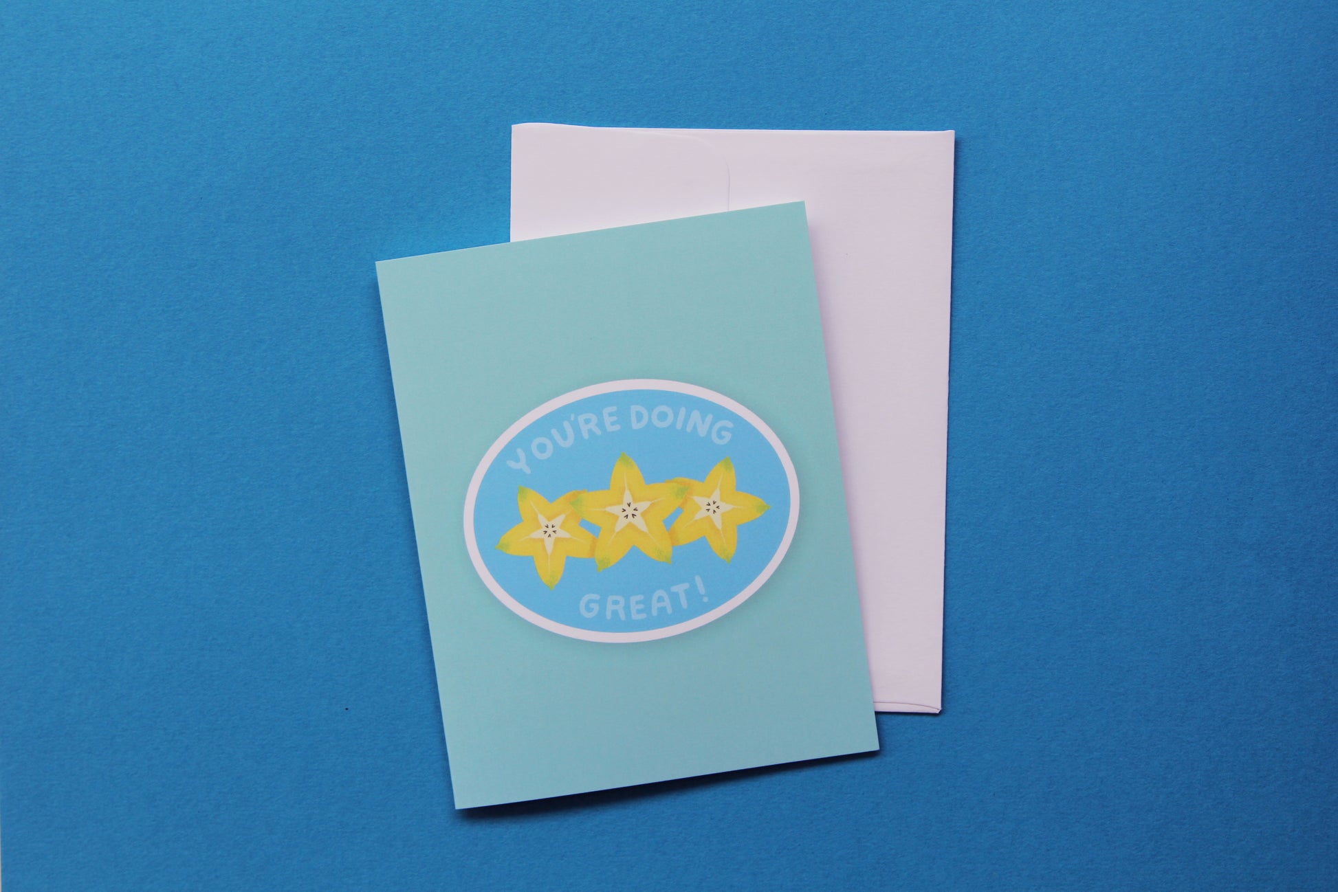 A photo of a blue greeting card with a sliced starfruit that says "You're doing great" and a white envelope on a blue background.