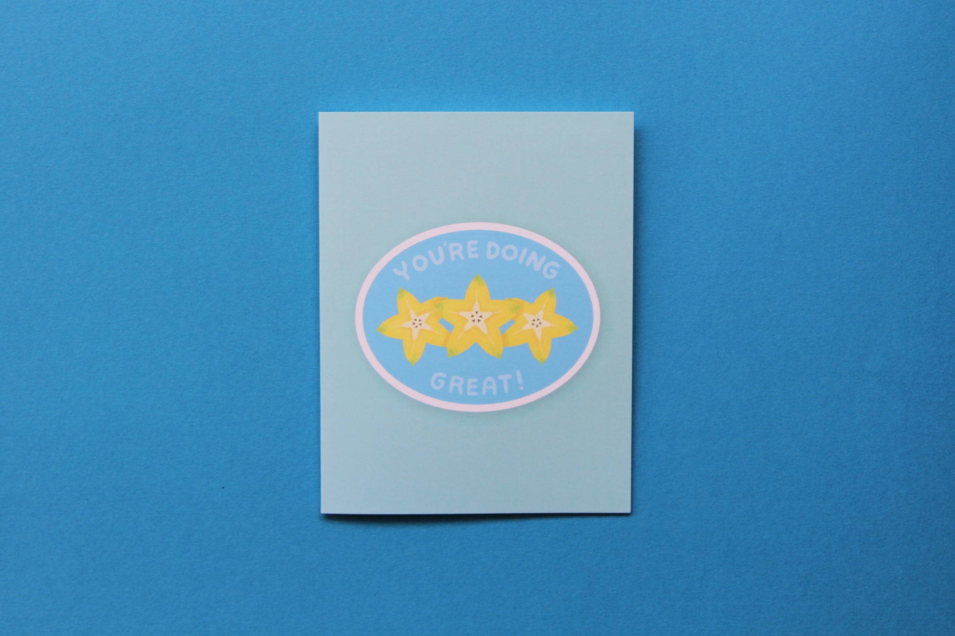 A photo of a blue greeting card with a sliced starfruit that says "You're doing great" on a blue background.