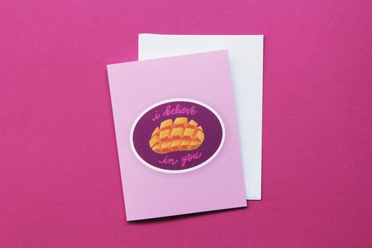 A photo of a pink greeting card with a sliced mango that says "I believe in you" and a white envelope on a pink background.
