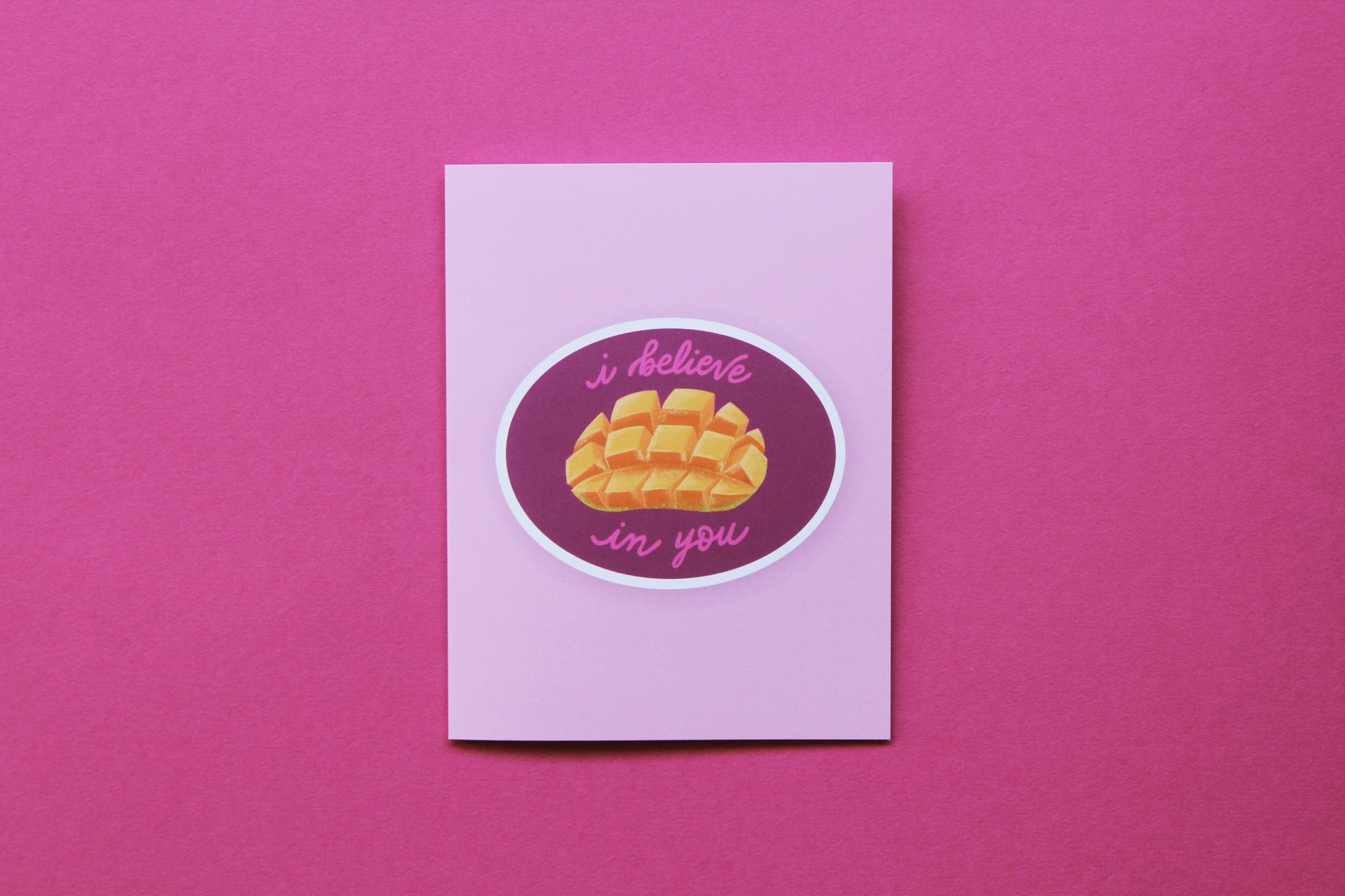 A photo of a pink greeting card with a sliced mango that says "I believe in you" on a pink background.