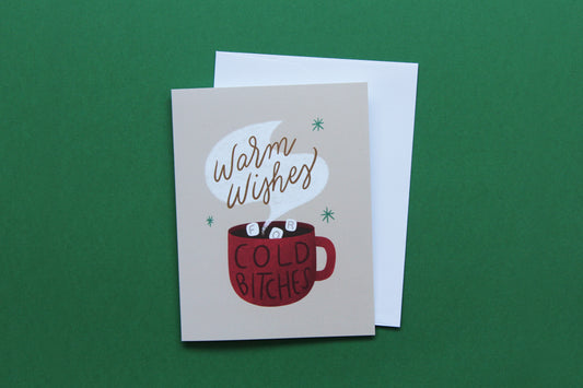 A photo of a tan greeting card with a cup of hot cocoa that says "Warm wishes for cold bitches" and a white envelope on a green background.