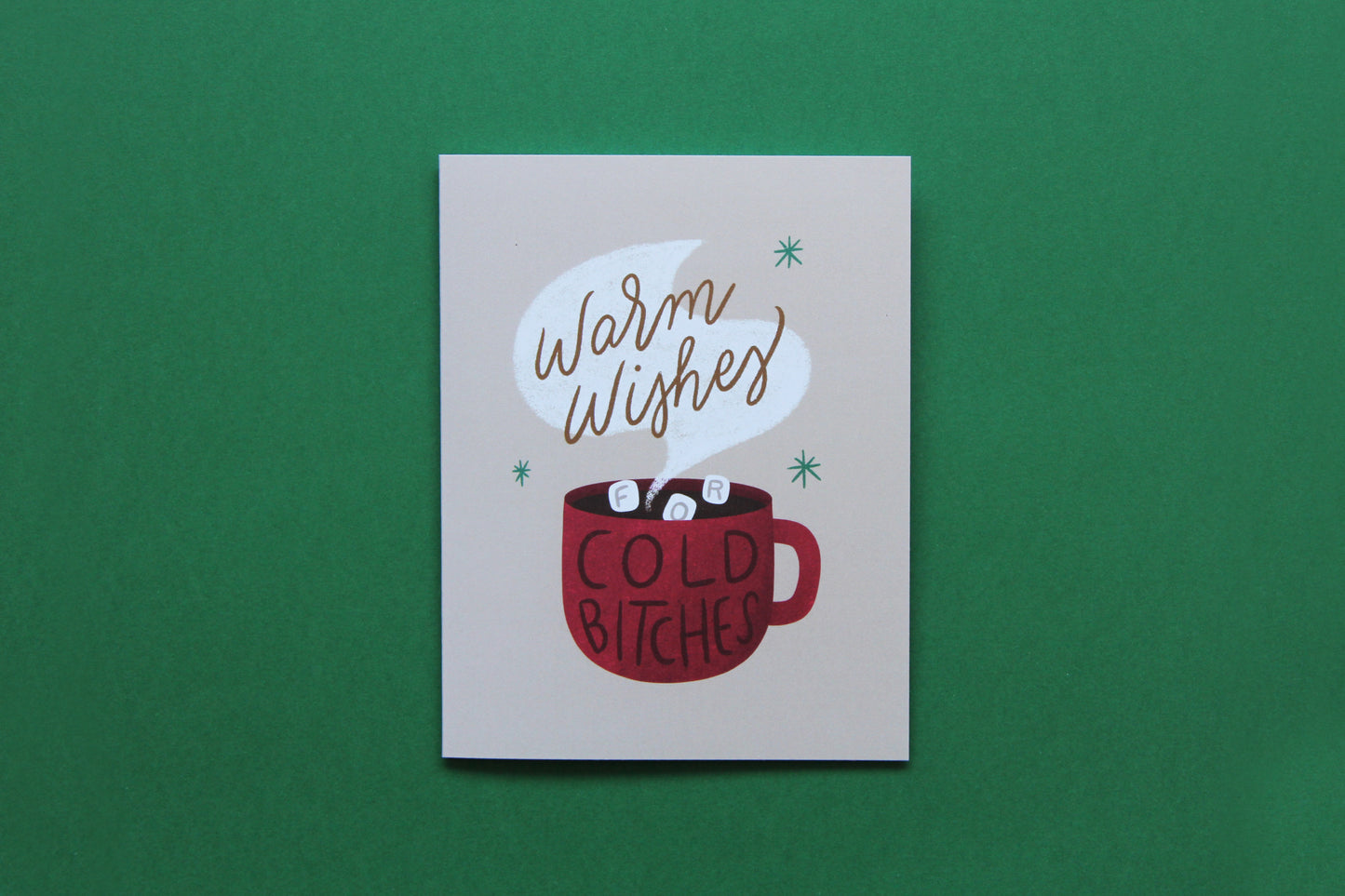 A photo of a tan greeting card with a cup of hot cocoa that says "Warm wishes for cold bitches" on a green background.