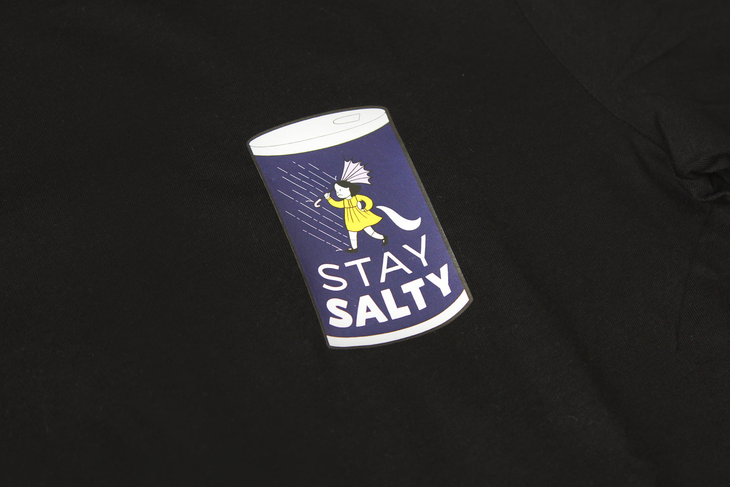 A closeup photo of a grumpy salt girl in a salt can that says "Stay Salty" printed on the right front chest of a black tshirt.