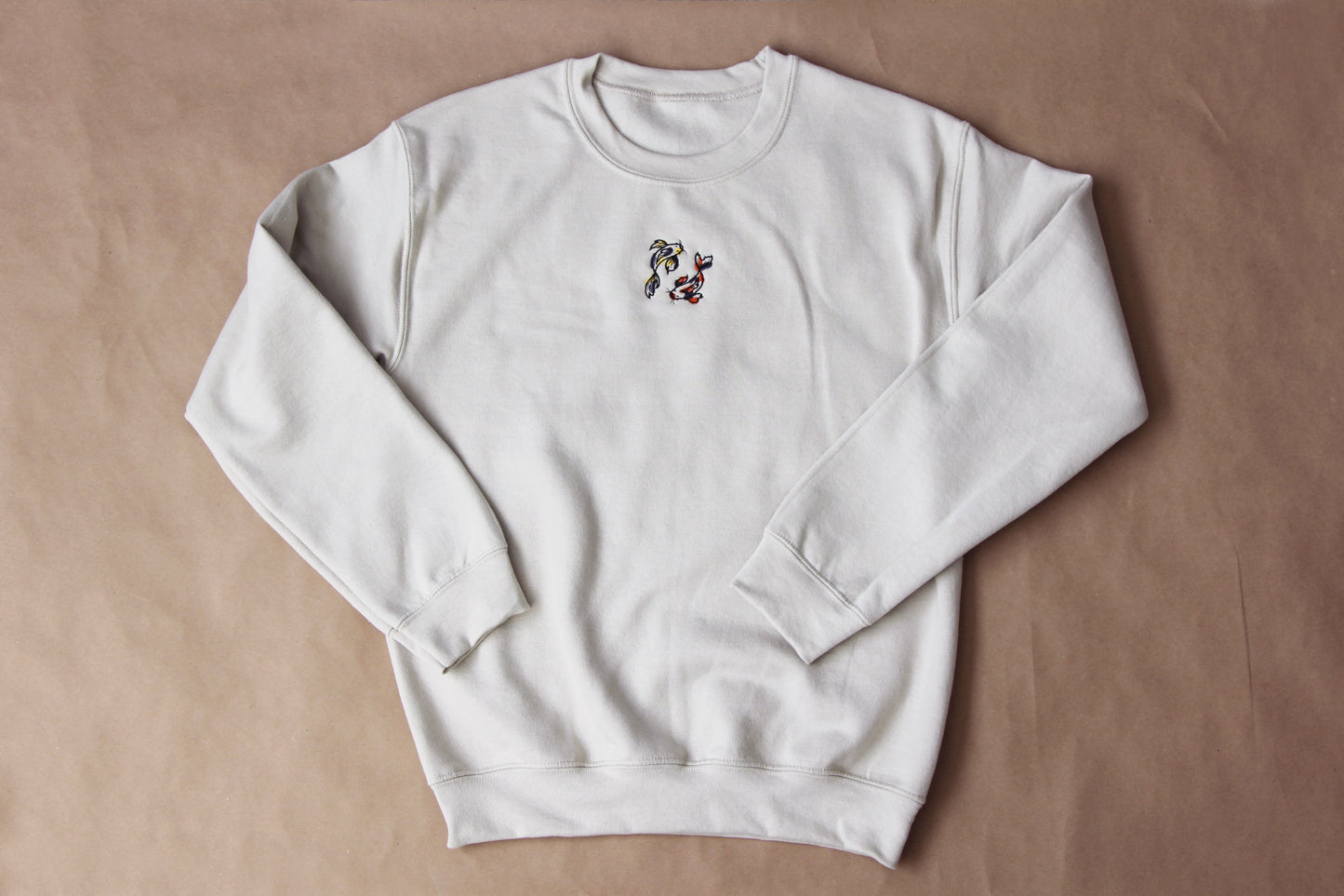 A photo of a cream crew neck sweatshirt with 2 koi fish embroidered on the front center over a tan background.