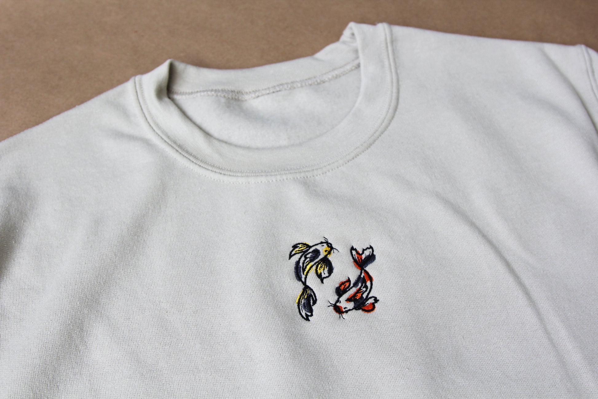 A closeup photo of 2 koi fish embroidered on the front center of a cream crew neck sweatshirt over a tan background.