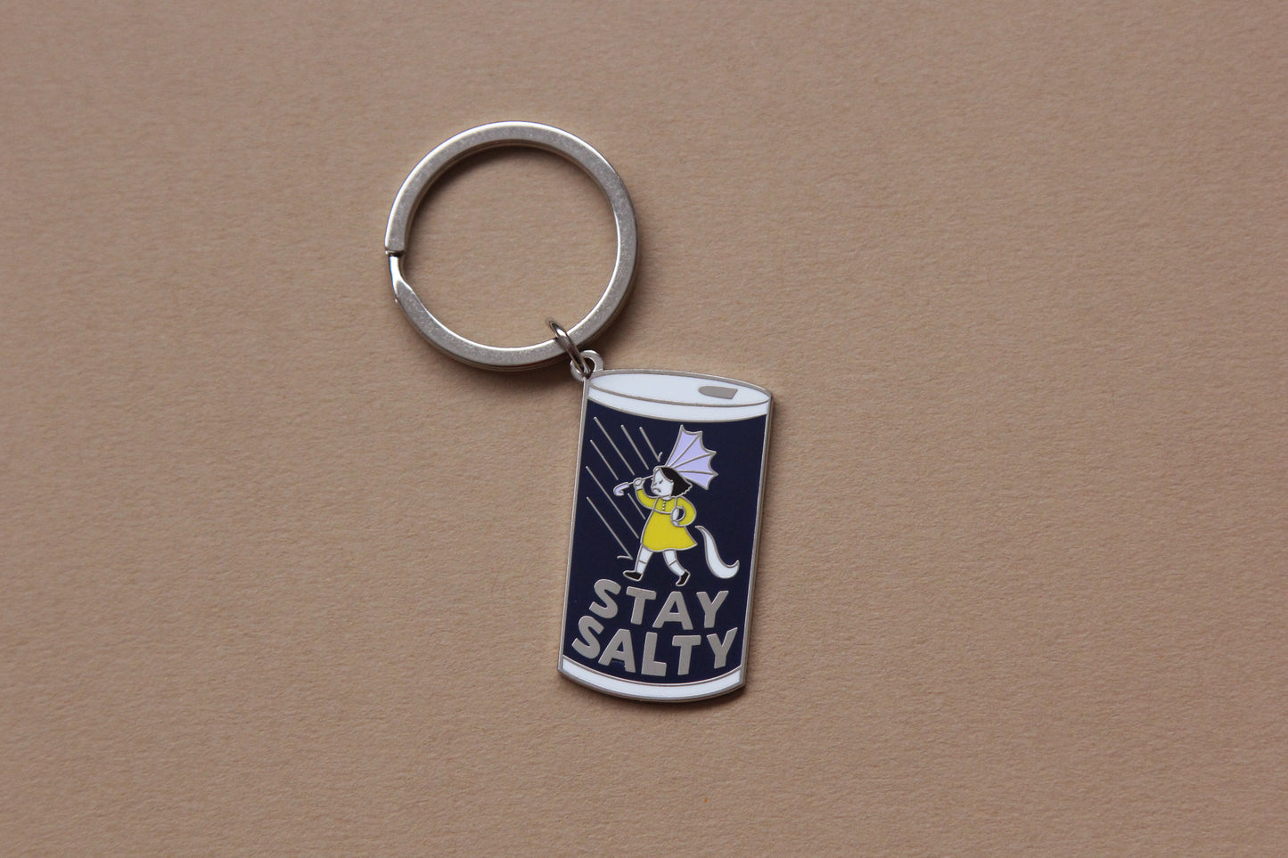 An enamel keychain showing a grumpy girl on a salt can that says "Stay Salty" over a tan background.