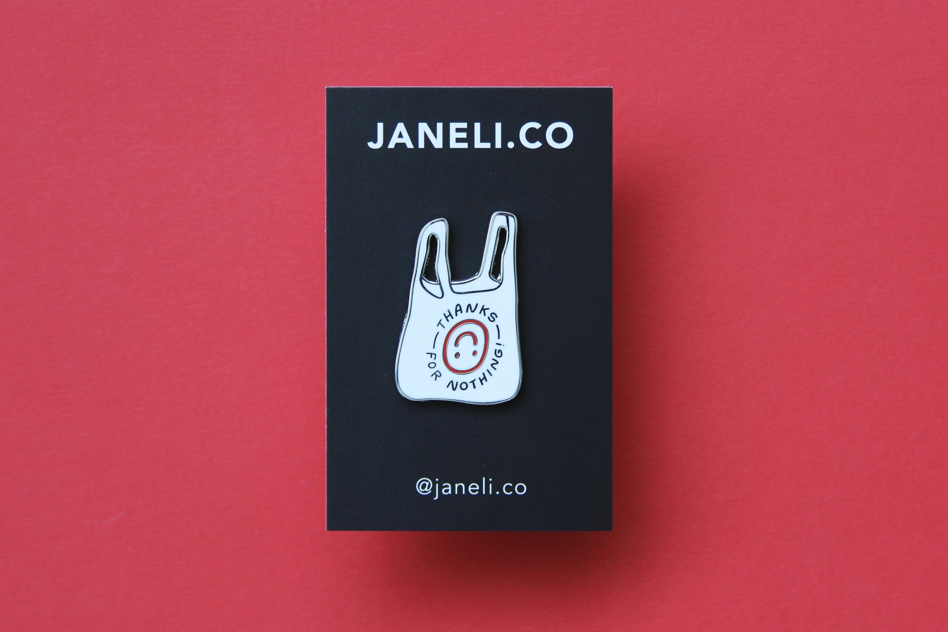 An enamel pin showing a plastic takeout bag that says "Thanks for nothing" with an upside down smiley face on a black JaneLi.Co backing card over a red background.
