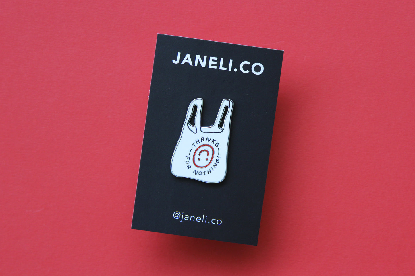 An enamel pin showing a plastic takeout bag that says "Thanks for nothing" with an upside down smiley face on a black JaneLi.Co backing card over a red background.