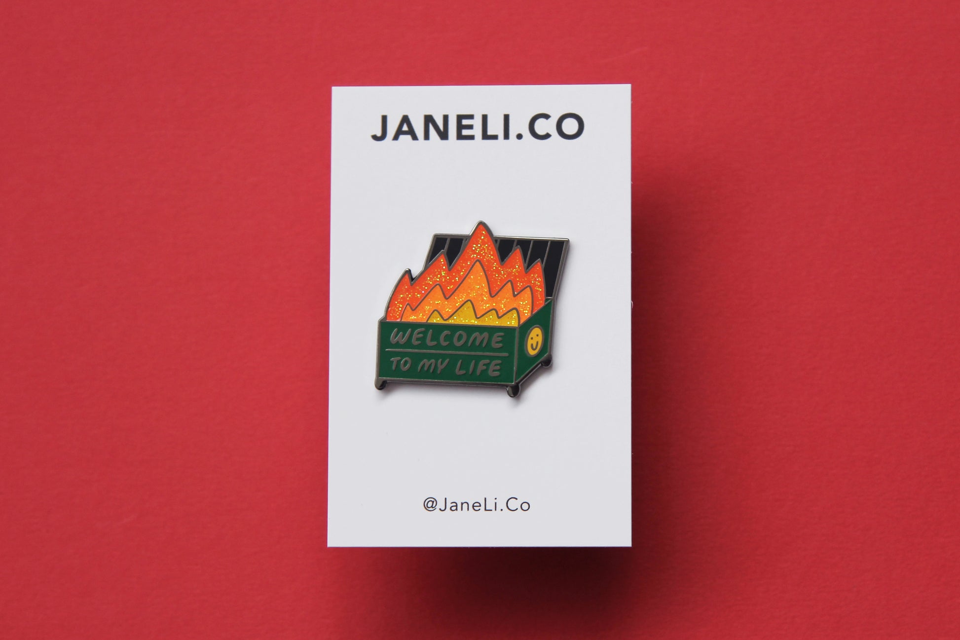 An enamel pin showing a dumpster with glittery flames that says "Welcome to my life" on a white JaneLi.Co backing card over a red background.