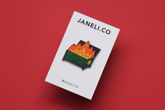 An enamel pin showing a dumpster with glittery flames that says "Welcome to my life" on a white JaneLi.Co backing card over a red background.