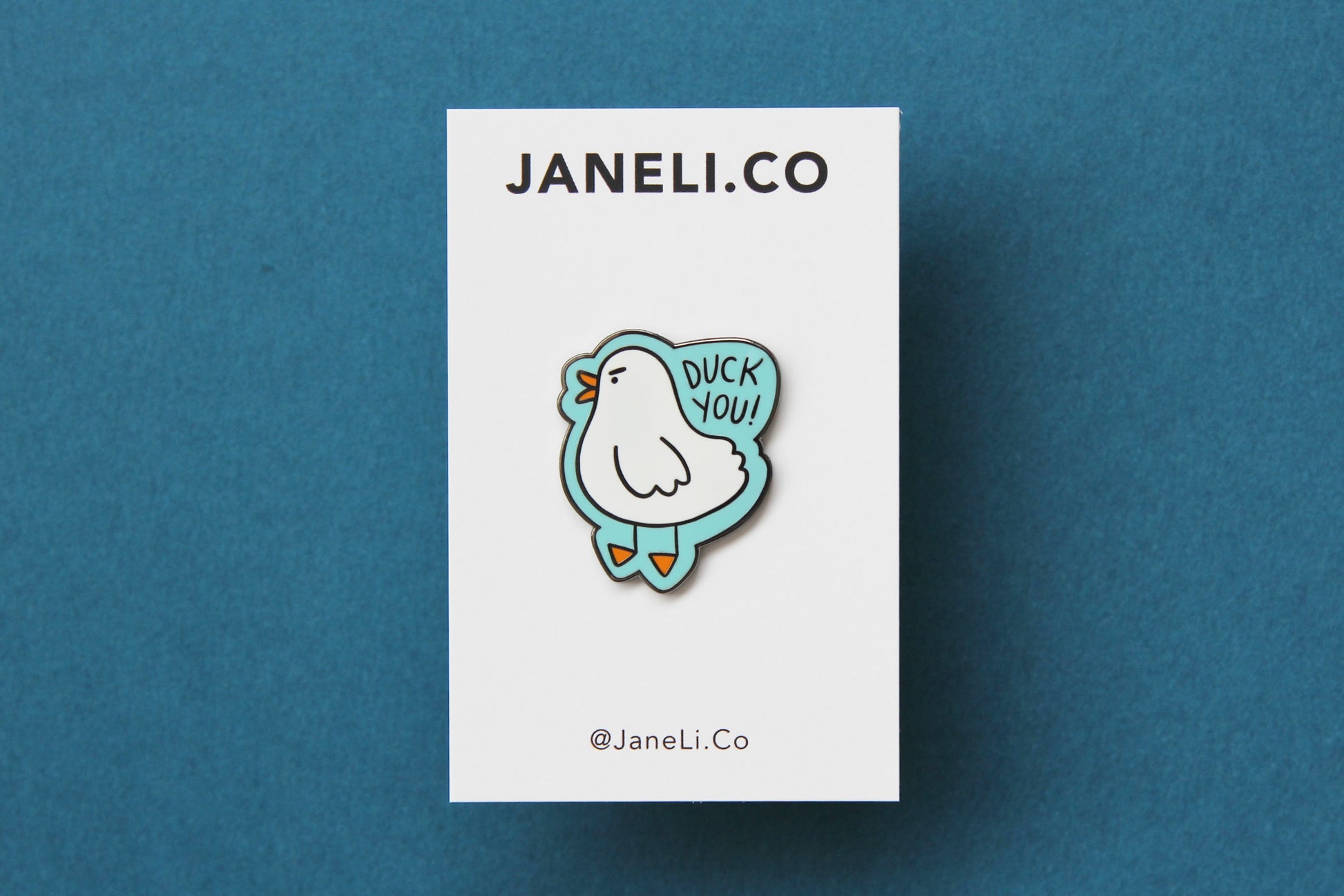 An enamel pin showing an angry white duck saying "Duck you!" on a white JaneLi.Co backing card over a teal background.