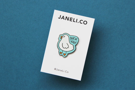 An enamel pin showing an angry white duck saying "Duck you!" on a white JaneLi.Co backing card over a teal background.