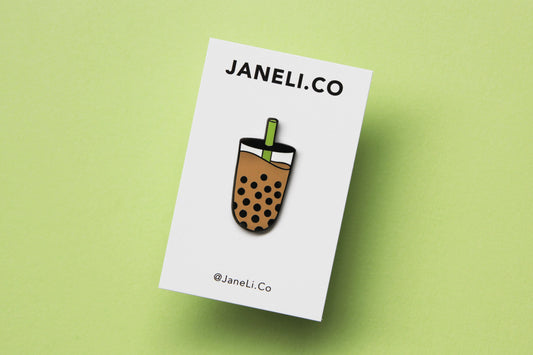 An enamel pin showing a cup of boba with a green straw on a white JaneLi.Co backing card over a green background.