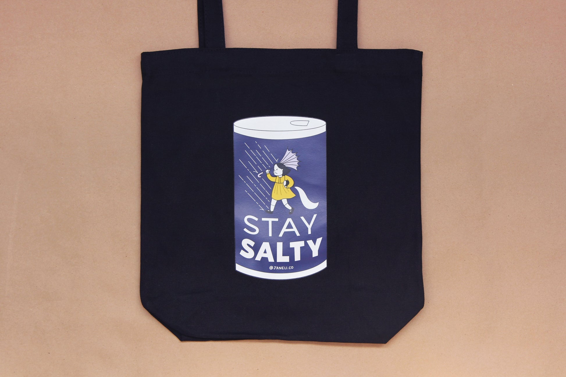 A photo of a black tote with a grumpy salt girl that says "Stay Salty" on it over a tan background.