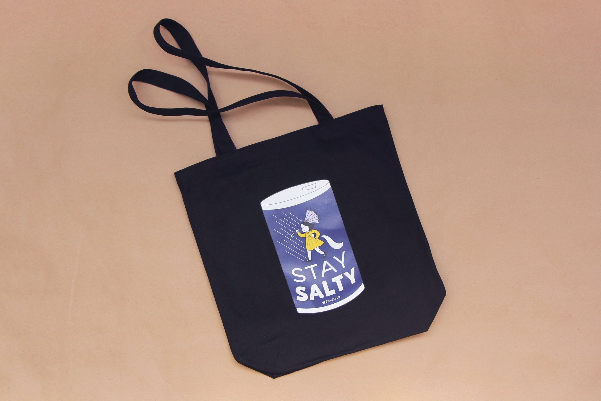 A photo of a black tote with a grumpy salt girl that says "Stay Salty" on it over a tan background.