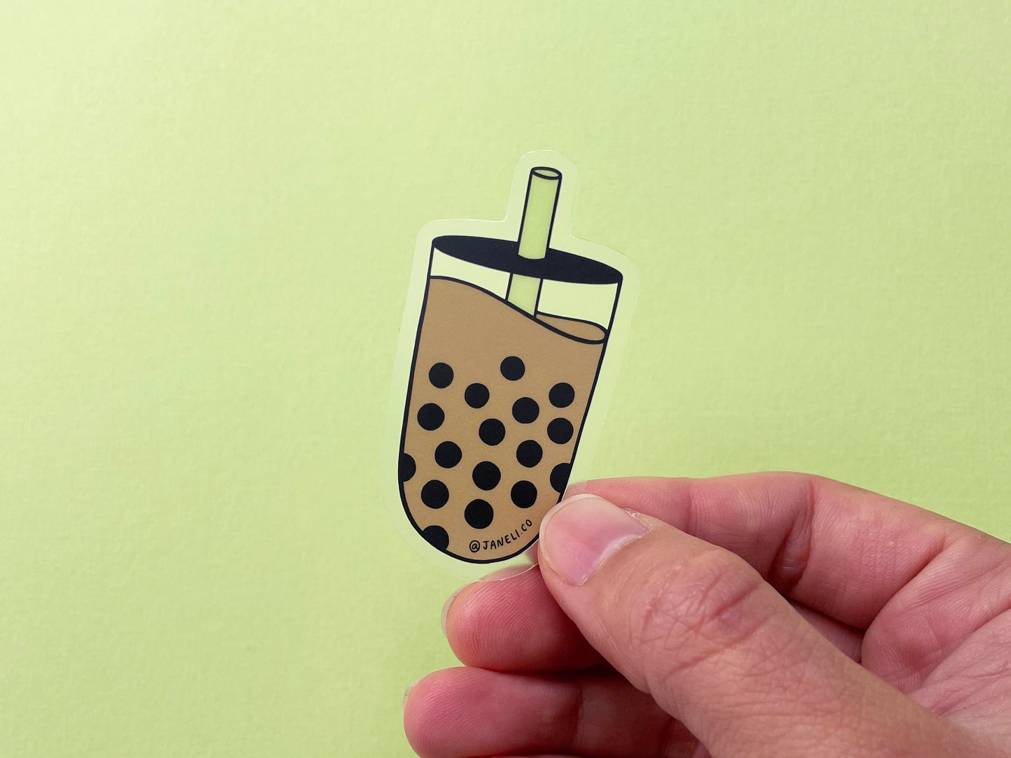 A hand holding a clear JaneLi.Co sticker showing a cup of a boba with a green straw over a green background.