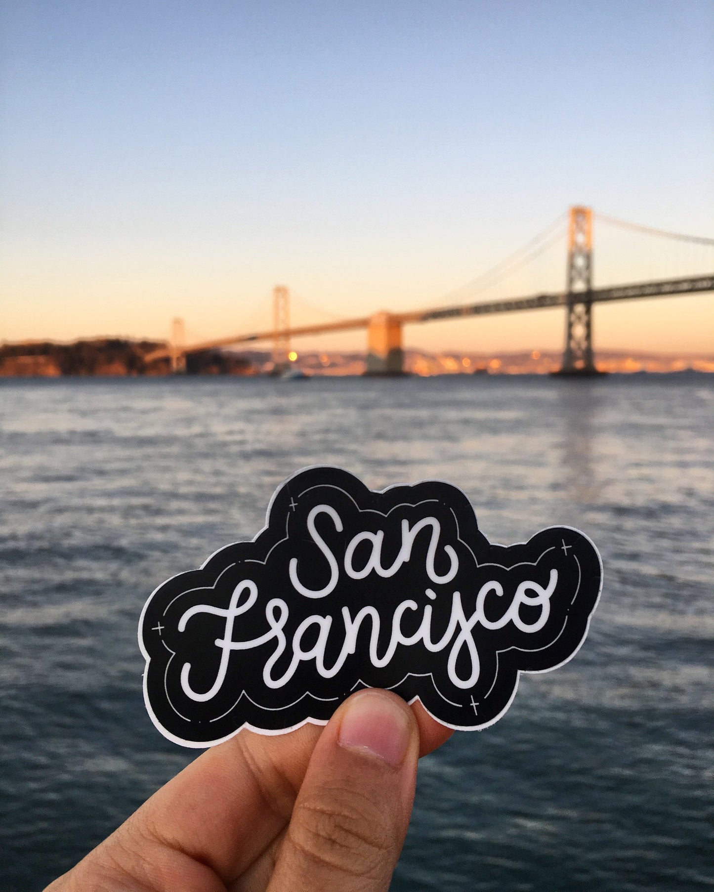 A hand holding a black and white JaneLi.Co sticker that says "San Francisco" in cursive lettering in front of the Bay Bridge in San Francisco, California.