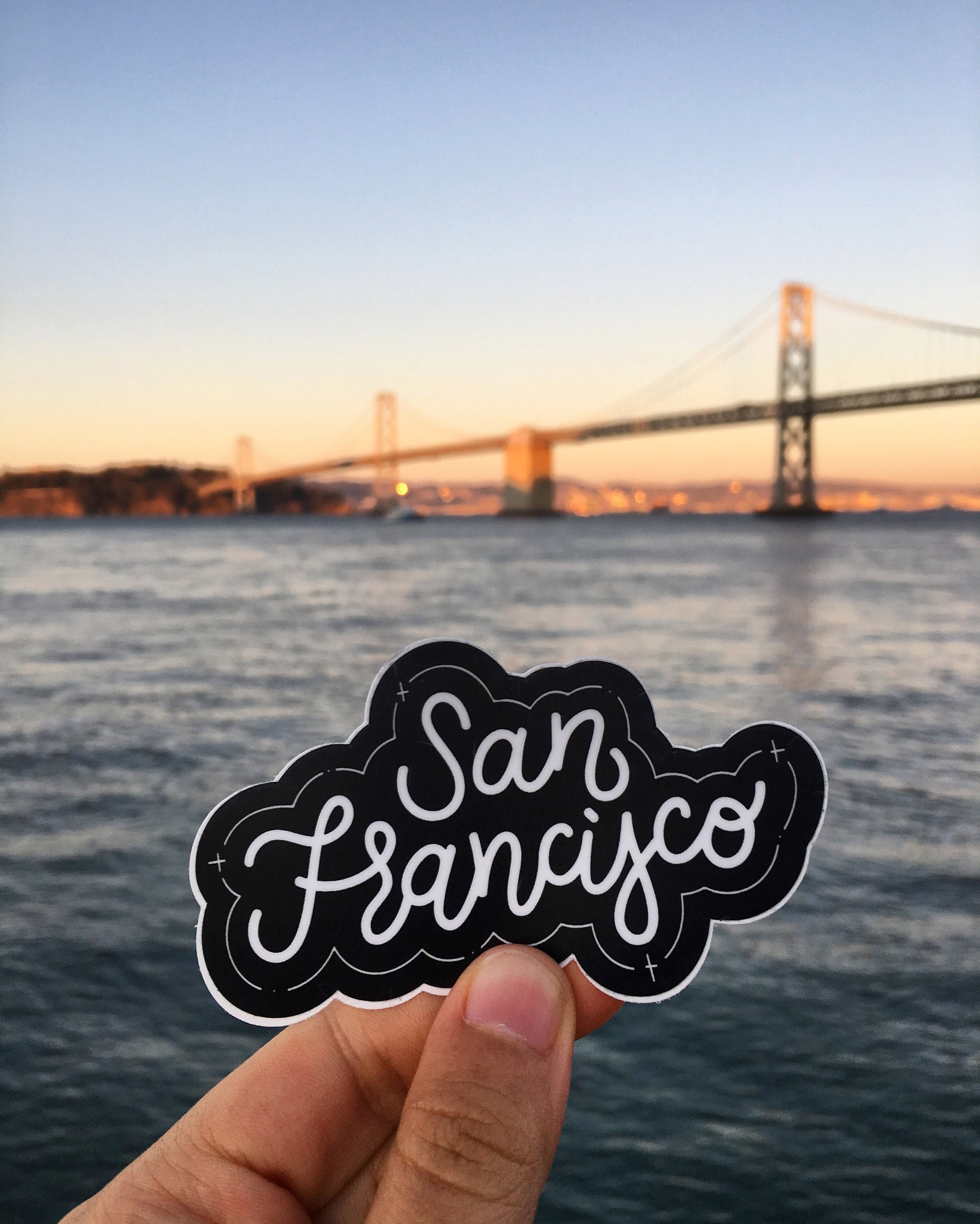 A hand holding a black and white JaneLi.Co sticker that says "San Francisco" in cursive lettering in front of the Bay Bridge in San Francisco, California.