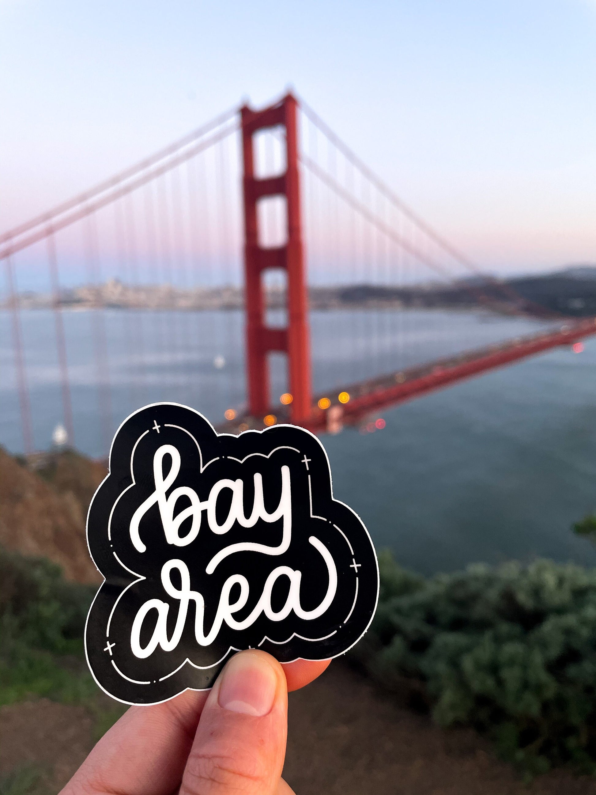 A hand holding a black and white JaneLi.Co sticker that says "Bay Area" in cursive lettering in front of the Golden Gate Bridge in San Francisco, California.