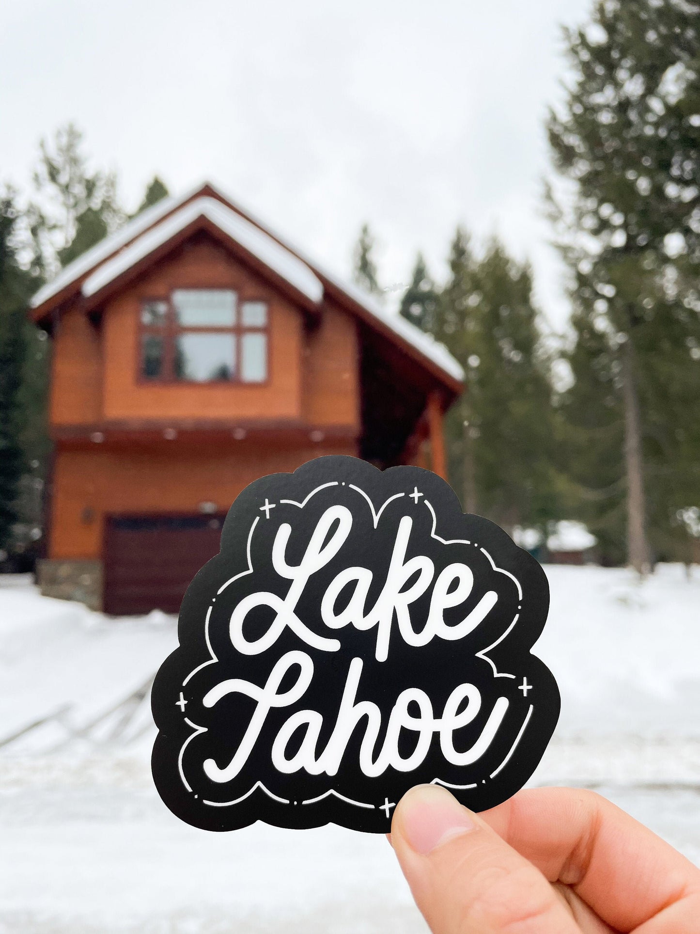 A hand holding a black and white JaneLi.Co sticker that says "Lake Tahoe" in cursive lettering in front of the snowy outdoors.