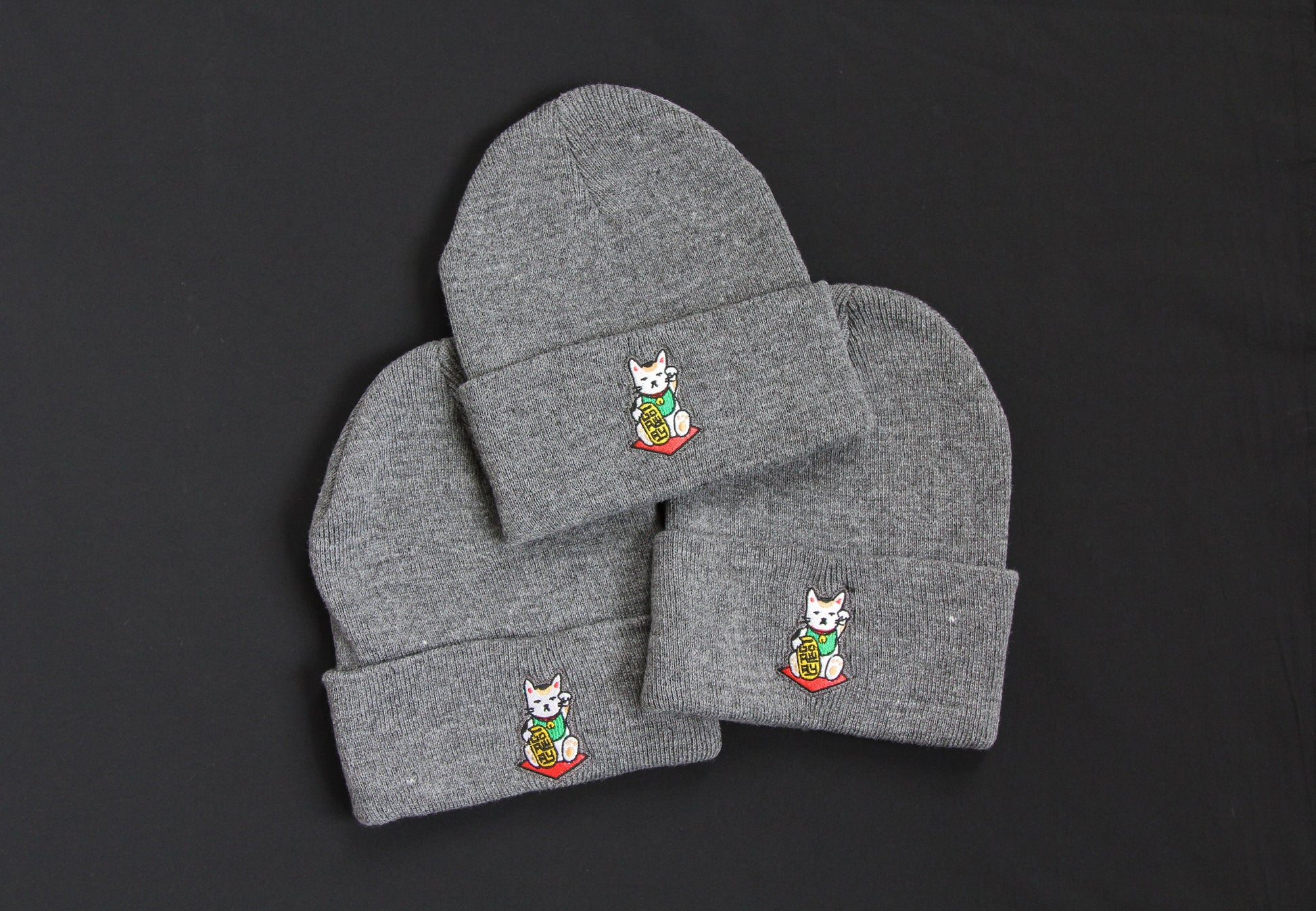 3 cuffed charcoal grey beanies with embroidered maneki neko cats holding gold bars that say "Go Away" on a black background. 