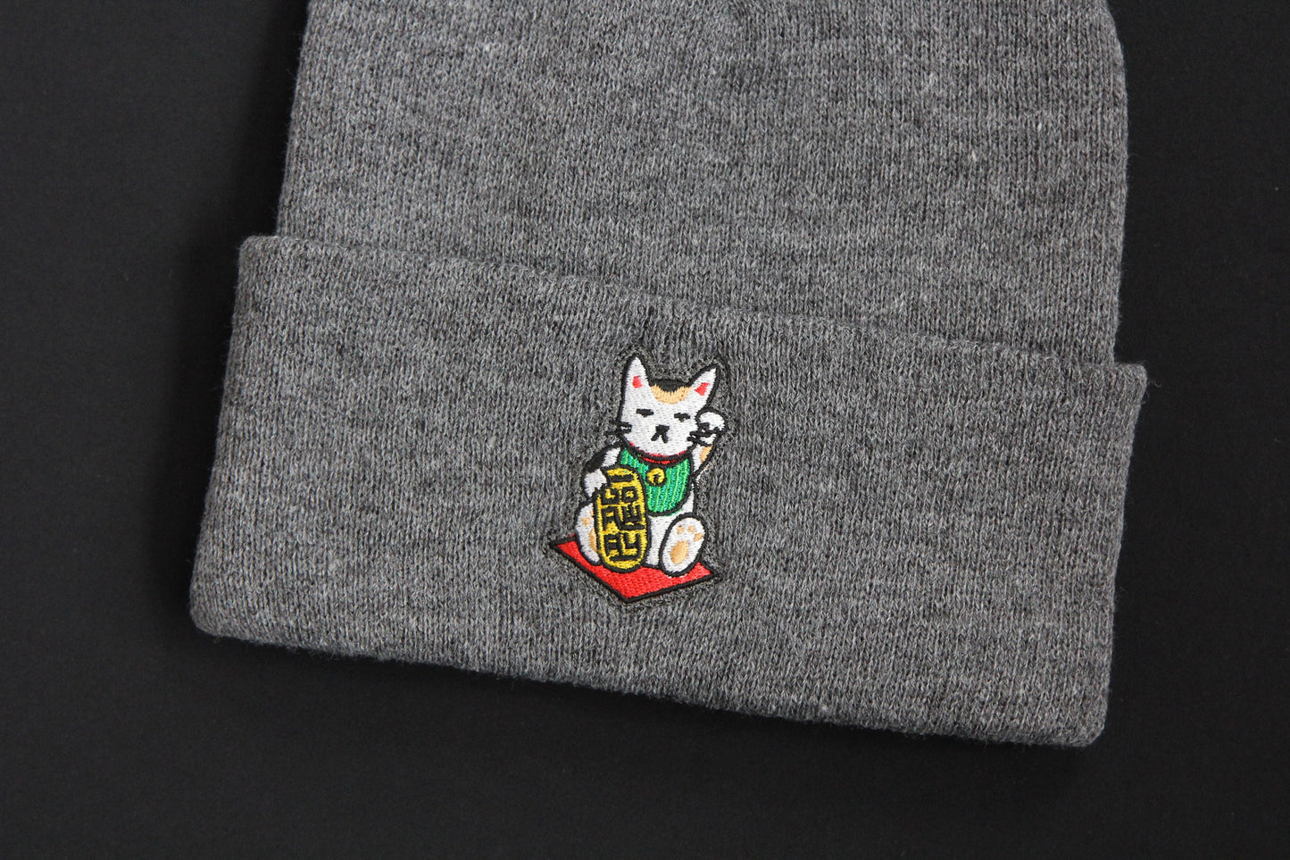 A close up of a cuffed charcoal grey beanie with an embroidered maneki neko cat holding a gold bar that says "Go Away" on a black background. 