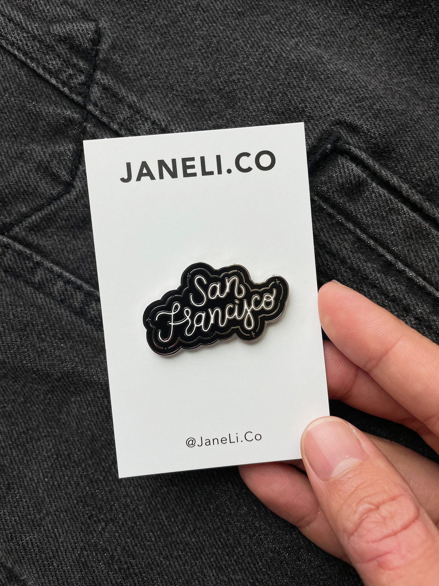A hand holding a black, white, and silver enamel pin that says "San Francisco" over a black denim jacket.
